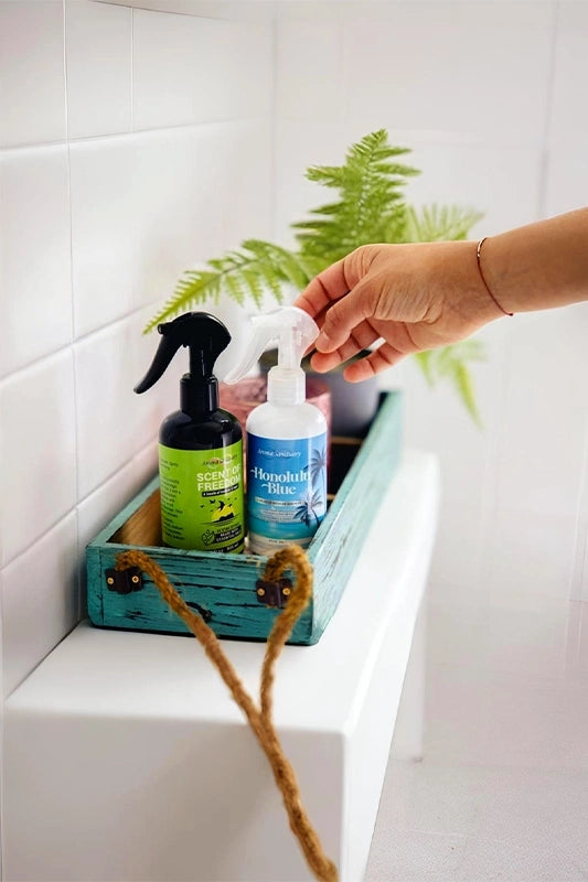 Scent of Freedom and Honolulu Blue plant-based room sprays by Aroma Sanctuary, placed in a wooden tray with a hand reaching for the bottle in a bathroom setting