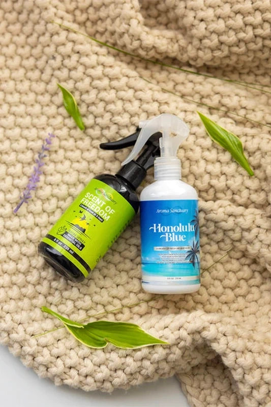 Scent of Freedom and Honolulu Blue plant-based room sprays by Aroma Sanctuary, placed on a beige knit blanket with green leaves and lavender sprigs